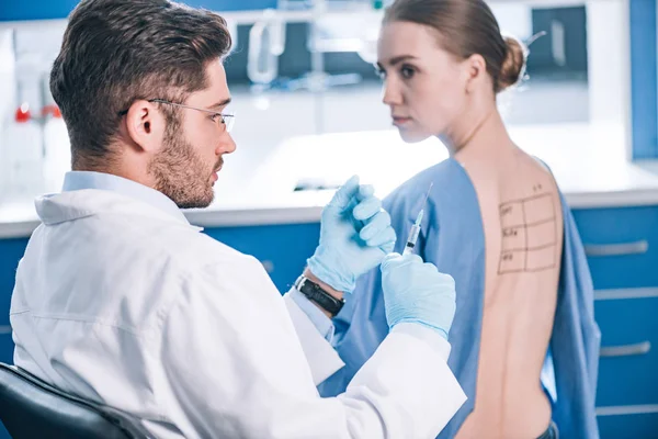 Selective focus of handsome allergist holding syringe near patient with letters on marked back — Stock Photo