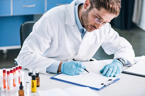 Selective focus of bearded immunologist in glasses holding pen near clipboard — Stock Photo