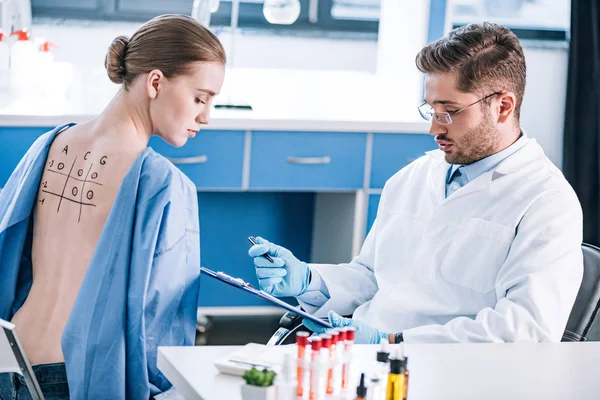 Selective focus of handsome allergist holding clipboard near woman with marked back — Stock Photo