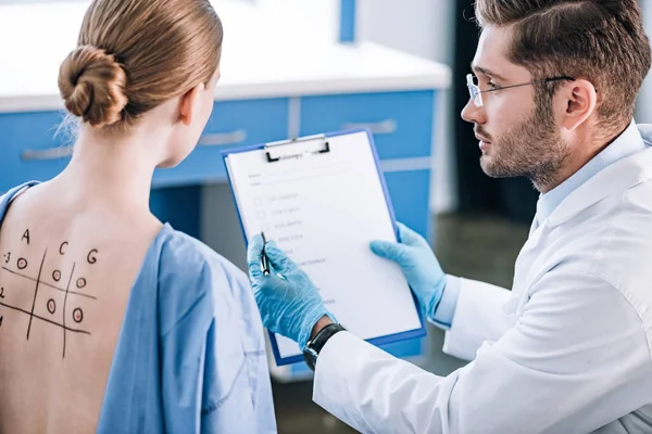 Foyer sélectif de beau immunologiste tenant presse-papiers près de la femme avec dos marqué — Photo de stock