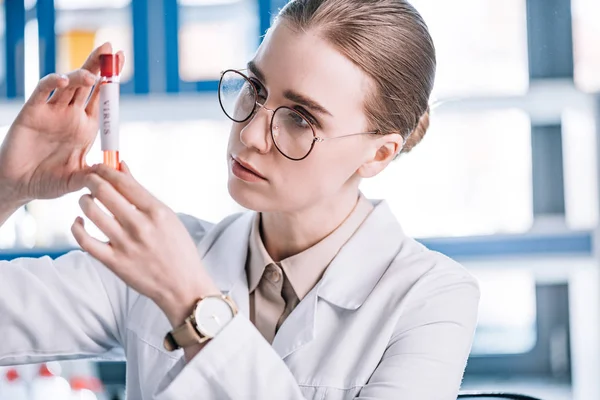 Selective focus of beautiful immunologist in glasses looking at test tube with virus letters — Stock Photo