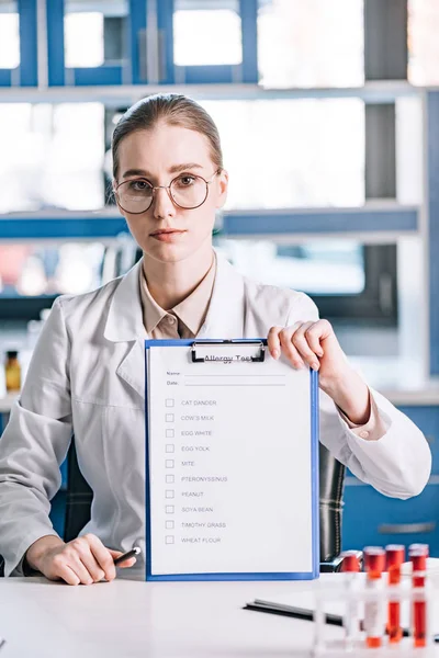 Attractive immunologist holding clipboard with checklist and pen near test tubes — Stock Photo