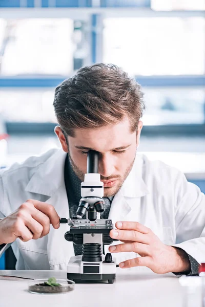 Foco seletivo do bioquímico barbudo no revestimento branco que olha através do microscópio no laboratório — Fotografia de Stock