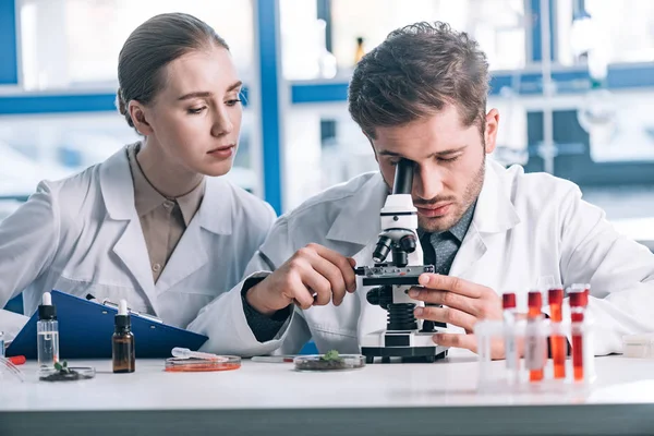 Messa a fuoco selettiva del biochimico che osserva attraverso il microscopio vicino al collega attraente — Foto stock