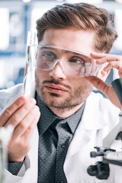 Selective focus of handsome biochemist touching goggles and looking a test tube — Stock Photo