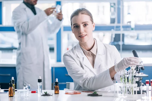 Selective focus of happy and attractive biochemist near microscope and plants in test tubes — Stock Photo