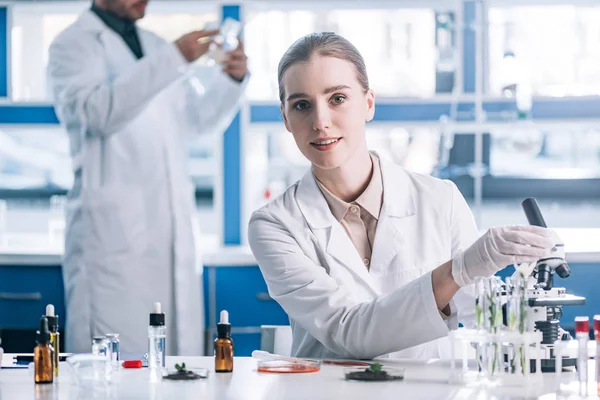 Foco seletivo de bioquímico feliz e atraente próximo ao microscópio e plantas em tubos de ensaio — Fotografia de Stock