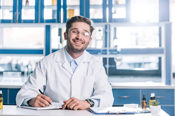 Hombre feliz en gafas y abrigo blanco sentado en el escritorio en la clínica - foto de stock