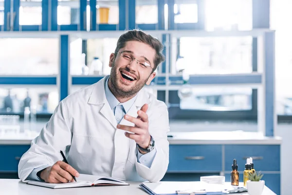 Cheerful doctor in glasses and white coat sitting at desk and gesturing in clinic — Stock Photo