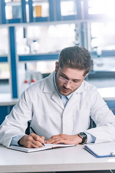 Schöner Allergologe schreibt in Notizbuch neben Klemmbrett auf Tisch — Stockfoto