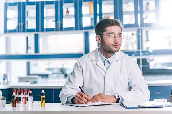 Pensive allergist writing in notebook near test tubes with samples — Stock Photo