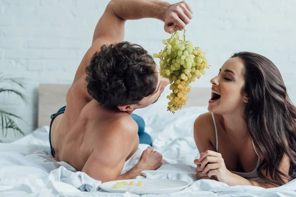 Hemdloser Mann hält Weintraube in der Nähe seiner Freundin, die mit offenem Mund und geschlossenen Augen liegt — Stockfoto