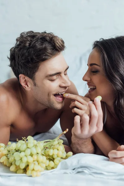 Счастливая молодая пара кормит друг друга виноградом, лежа на кровати — стоковое фото