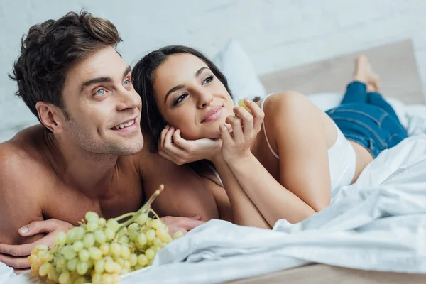 Счастливая молодая пара смотрит в сторону, лежа на кровати рядом с гроздью винограда — стоковое фото