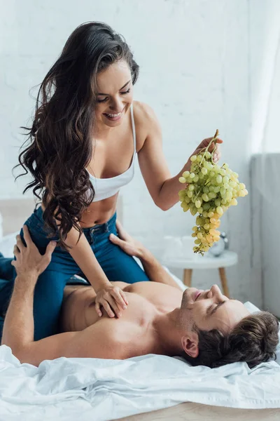 Sexy, smiling woman sitting on men and holding bunch of grapes — Stock Photo