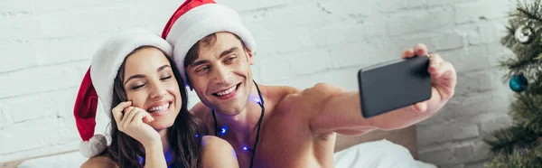 Panoramic shot of cheerful couple in santa hats taking selfie on smartphone in bed — Stock Photo