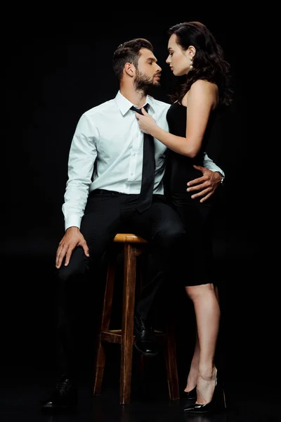 Beautiful woman looking at bearded man sitting on chair isolated on black — Stock Photo