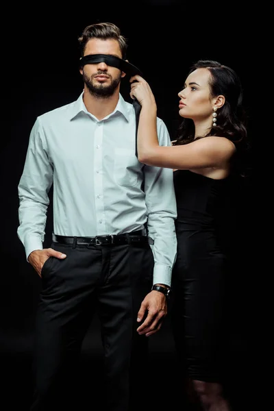 Attraktive Frau mit Augenbinde auf Mann stehend mit Hand in Tasche isoliert auf schwarz — Stockfoto