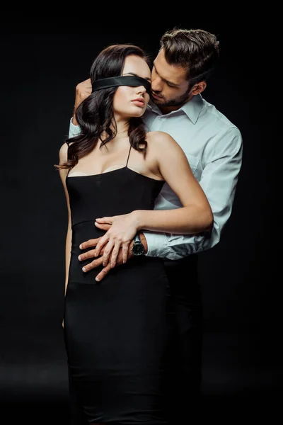 Handsome man looking at blindfolded young woman on black — Stock Photo