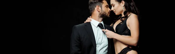 Panoramic shot of woman in sexy lingerie touching tie of bearded boyfriend isolated on black — Stock Photo