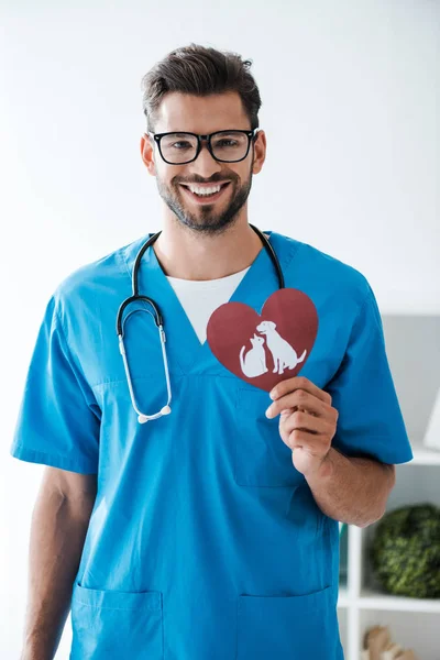 Handsome veterinarian smiling at camera while presenting paper cut heart with dog and cat symbols — Stock Photo