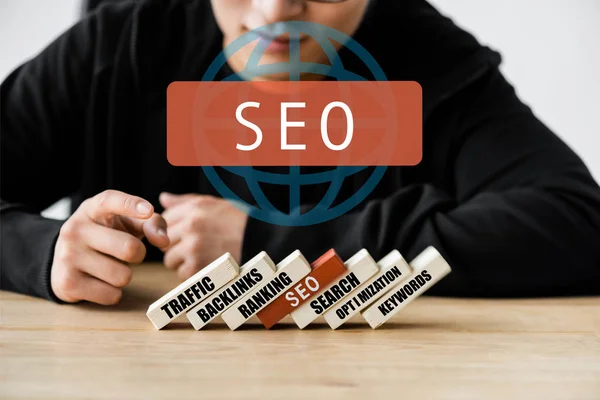 Cropped view of seo manager pointing with finger and sitting near wooden rectangles with illustration of concept words of seo — Stock Photo