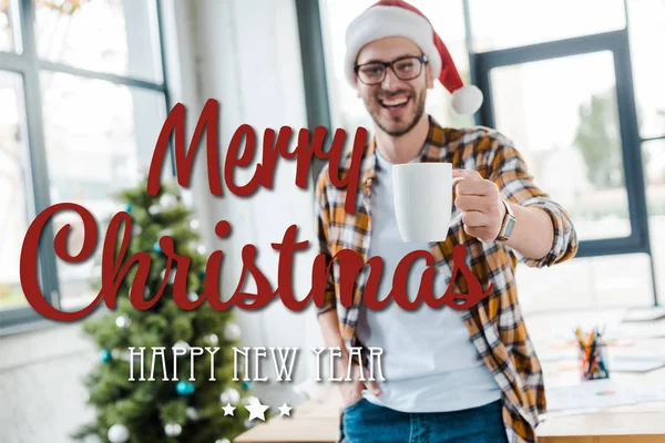 Селективное внимание счастливого бородатого мужчины в шляпе Санты, держащего чашку возле рождественской елки в офисе с весёлым Рождеством и счастливой новогодней иллюстрацией — стоковое фото