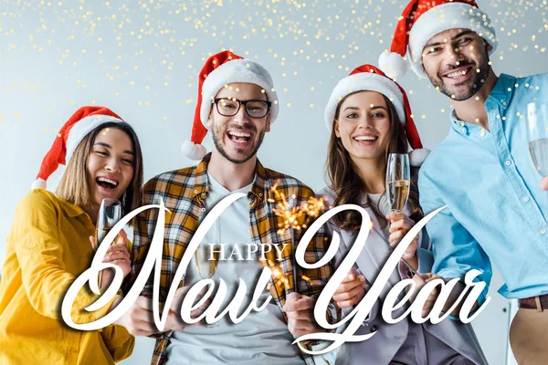 Selective focus of happy businessmen and multicultural businesswomen holding sparklers and champagne glasses in office with happy new year illustration — Stock Photo