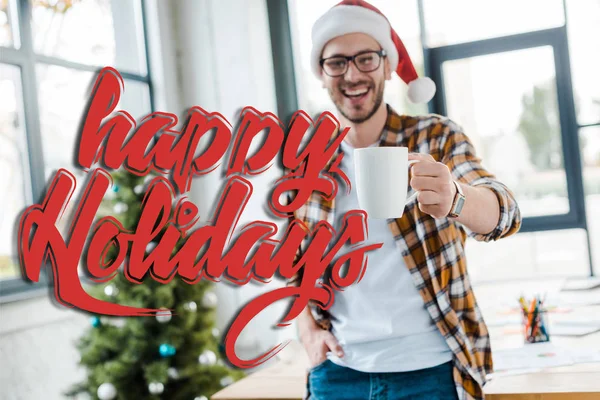 Foco seletivo de homem barbudo feliz em santa chapéu segurando copo perto de árvore de natal no escritório com feliz ilustração feriados — Fotografia de Stock