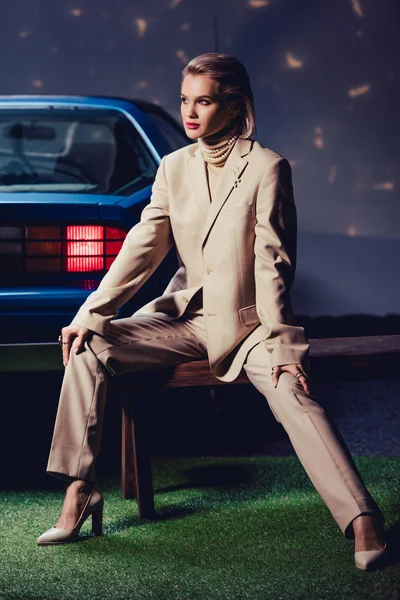 Привлекательная и стильная женщина в костюме сидит на деревянной скамейке возле ретро-машины — стоковое фото