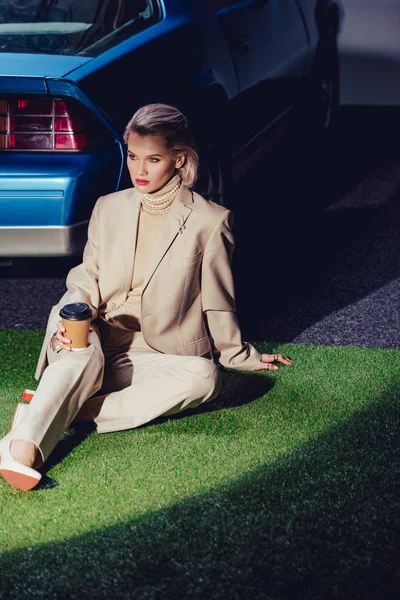 Attrayant et élégant femme en costume assis près de voiture rétro et tenant tasse en papier — Photo de stock