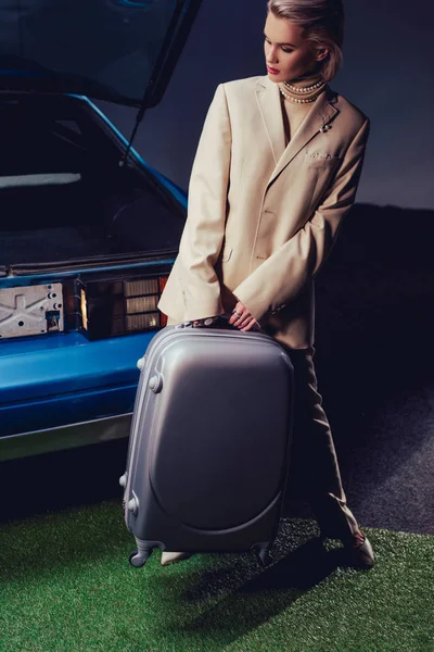 Приваблива і стильна жінка в костюмі тримає валізу і стоїть біля ретро-автомобіля — стокове фото