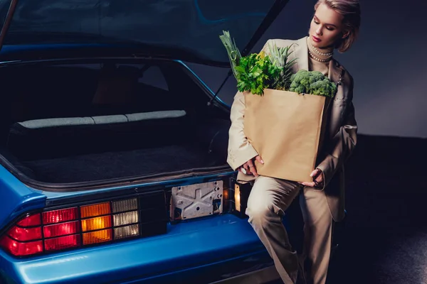 Attractive and stylish woman in suit standing near retro car and holding paper bag with food — Stock Photo