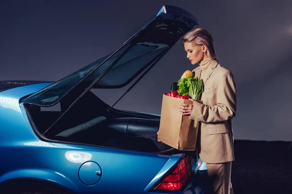 Side view of attractive and stylish woman in suit standing near retro car and holding paper bag with food — Stock Photo