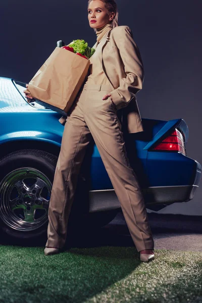Attractive and stylish woman in suit standing near retro car and holding paper bag with food — Stock Photo