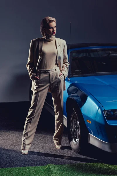 Mujer atractiva y elegante en traje con las manos en los bolsillos de pie cerca de coche retro - foto de stock