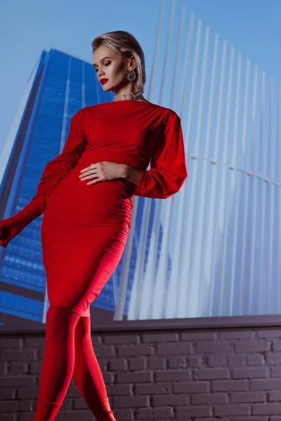 Vista de ángulo bajo de la mujer atractiva y elegante en vestido rojo en el fondo de la ciudad - foto de stock