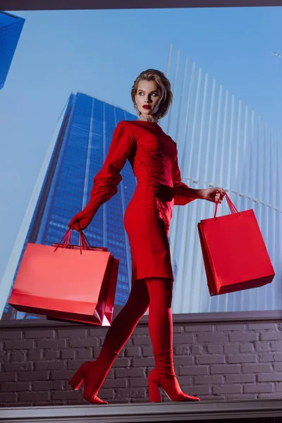 Baixo ângulo vista de mulher atraente e elegante em vestido vermelho segurando sacos de compras no fundo da cidade — Fotografia de Stock