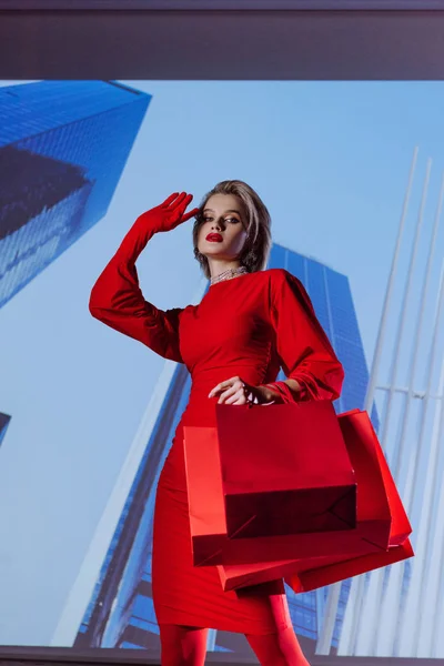 Vista basso angolo di donna attraente ed elegante in abito rosso con borse della spesa sullo sfondo della città — Foto stock
