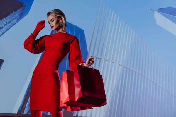 Vue à angle bas de femme attrayante et élégante en robe rouge tenant des sacs à provisions sur fond de ville — Photo de stock