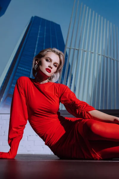 Mulher atraente e elegante em vestido vermelho sentado no fundo da cidade — Fotografia de Stock