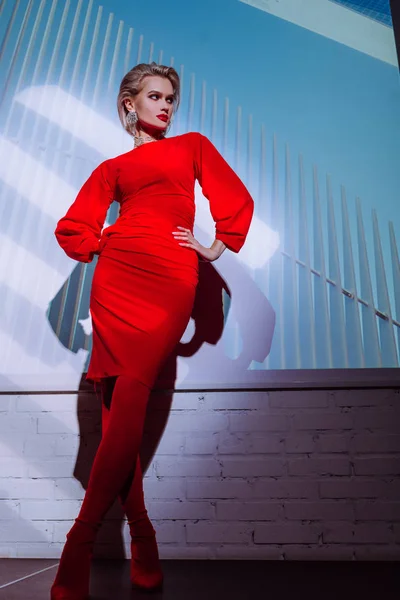 Vista de ángulo bajo de la mujer atractiva y elegante en vestido rojo con las manos en las caderas en el fondo de la ciudad - foto de stock