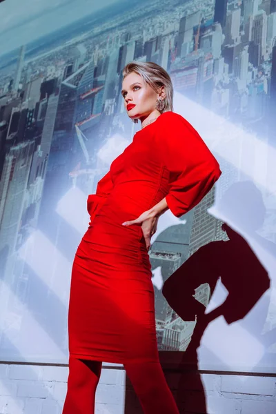 Low angle view of attractive and stylish woman with hand on hip in red dress on city background — Stock Photo