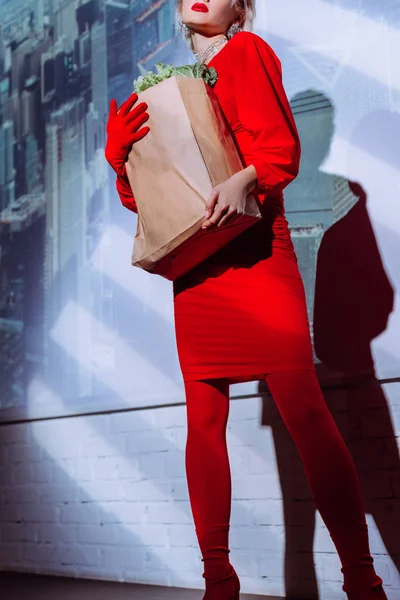 Vue recadrée de femme élégante en robe rouge tenant sac en papier avec de la nourriture sur le fond de la ville — Photo de stock