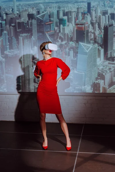 Stylish woman with hands on hips in red dress and virtual reality headset on city background — Stock Photo