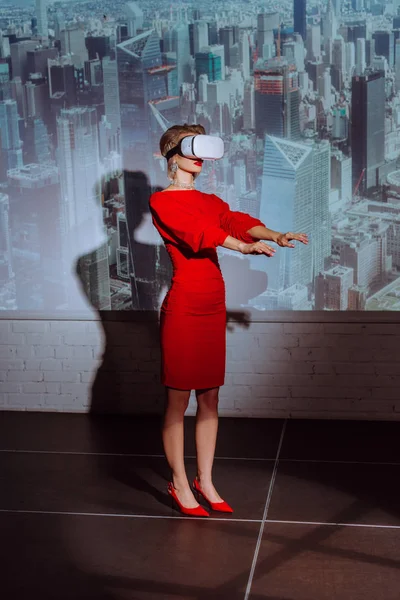Mujer elegante con las manos extendidas en vestido rojo y auriculares de realidad virtual en el fondo de la ciudad - foto de stock