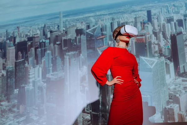 Vista basso angolo di donna elegante con le mani sui fianchi in abito rosso e auricolare realtà virtuale sullo sfondo della città — Foto stock