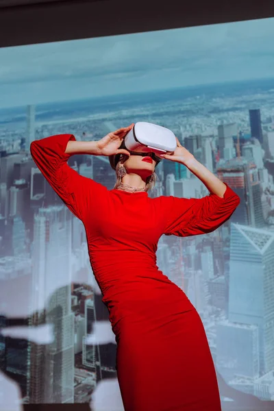 Low angle view of stylish woman in red dress and virtual reality headset on city background — Stock Photo