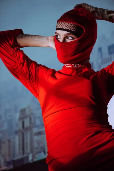 Low angle view of stylish woman in red dress and balaclava on city background — Stock Photo