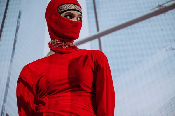 Baixo ângulo vista de mulher elegante em vestido vermelho e balaclava no fundo da cidade — Fotografia de Stock